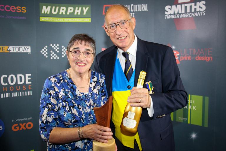 Rik and Marian Ludlow smiling holding award trophy and prosecco bottle. 